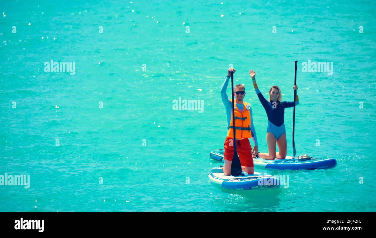 Mann und Frau rudern auf Stand-Up-Paddle-Brettern. Junger Mann in Badeanzug und Schwimmweste und junge Frau, die auf einem Stand-Up-Paddle-Board winkt. Sommer raus Stockfoto