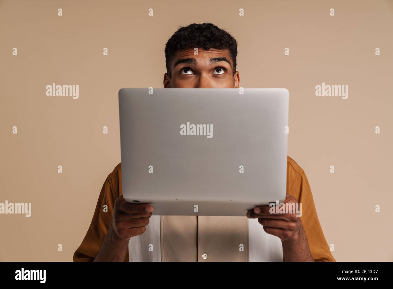 Ein junger, gutaussehender afrikaner, der hinter einem Laptop nach oben blickte, während er über einem isolierten beigen Hintergrund stand Stockfoto