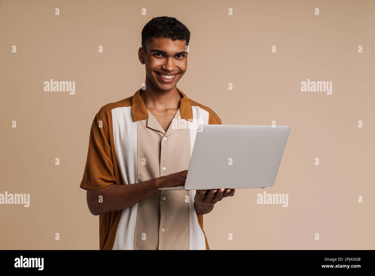 Ein junger, gut aussehender afrikaner mit einem durchdringenden Laptop und Blick in die Kamera, während er über einem isolierten beigen Hintergrund stand Stockfoto