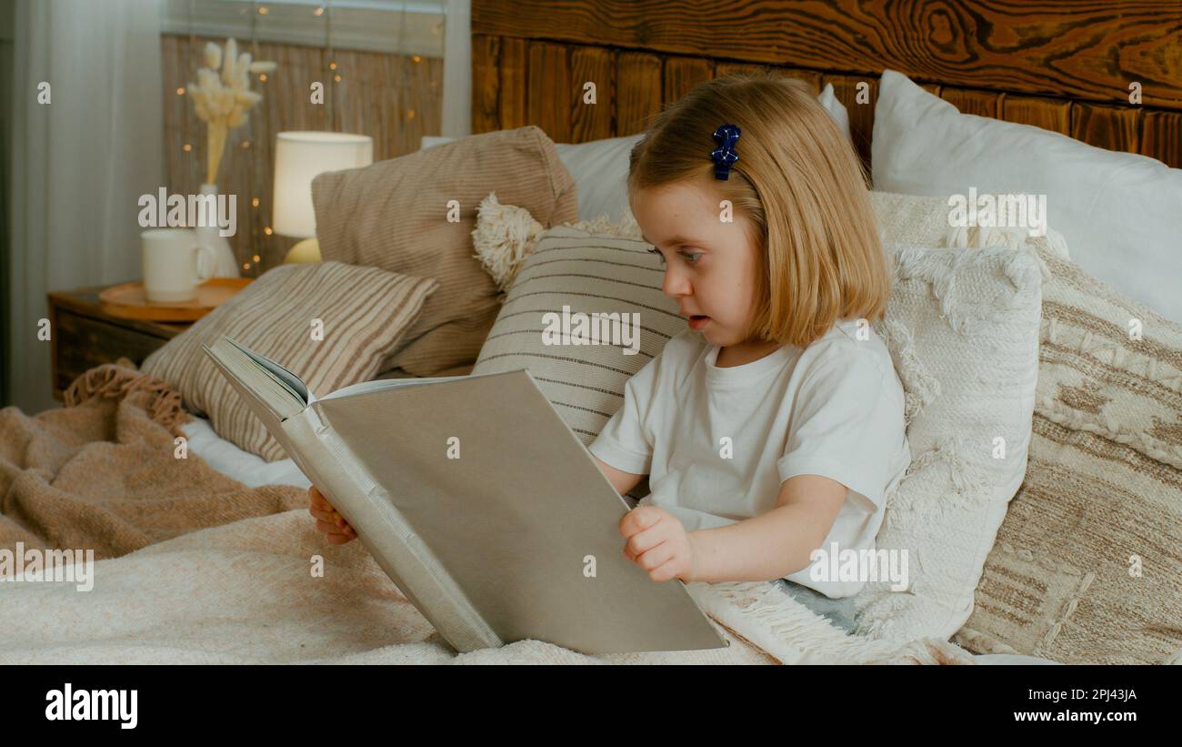 Süße kleine, 5s-jährige, alleinstehende, kaukasische Tochter, die in ein Buch schaut Kind Vorschulkind Baby sitzt im Bett und sieht sich Bilder in einem Copybook an und lernt Stockfoto