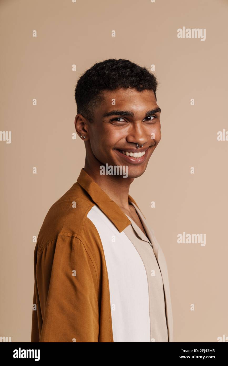 Porträt eines jungen, gutaussehenden, lächelnden afrikanischen Mannes mit einem Piercing, der in die Kamera schaut, während er mit seiner Seite über einem isolierten beigen Hintergrund steht Stockfoto