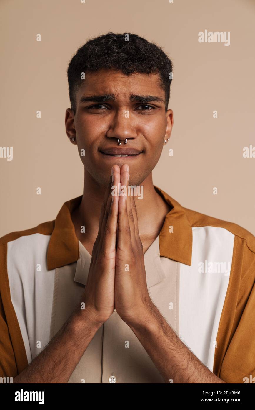 Junger, attraktiver, angespannter afrikaner mit geschlossenen Augen, der mit gefalteten Händen betet, während er über einem isolierten beigen Hintergrund steht Stockfoto