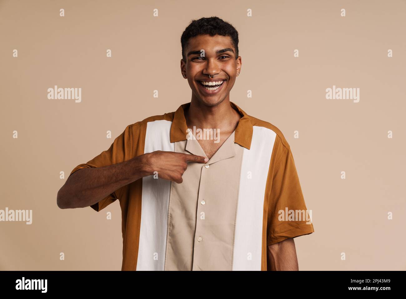 Ein junger, gutaussehender, glücklicher afrikaner mit Piercing, der auf sich selbst zeigt und in die Kamera schaut, während er über einem isolierten beigen Hintergrund steht Stockfoto