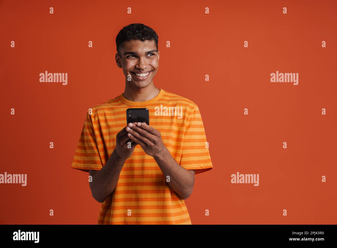 Ein junger, gut aussehender afrikaner mit Piercings, der sein Handy hielt und zur Seite schaute, während er über einem isolierten orangefarbenen Hintergrund stand Stockfoto