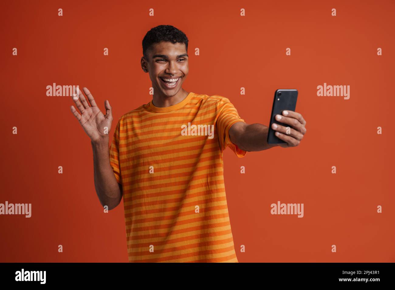 Ein junger, gut aussehender, glücklicher afrikaner, der pierdelnd winkt und Selfie am Telefon macht, während er über einem isolierten orangefarbenen Hintergrund steht Stockfoto