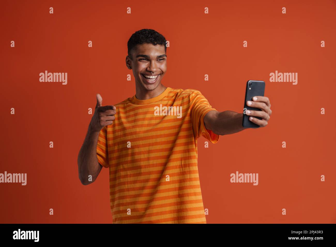 Junger, gutaussehender, glücklicher afrikanischer Mann mit Piercing, der eine Gesten mit Pistole macht und Selfie am Telefon macht, während er über einem isolierten orangefarbenen Hintergrund steht Stockfoto