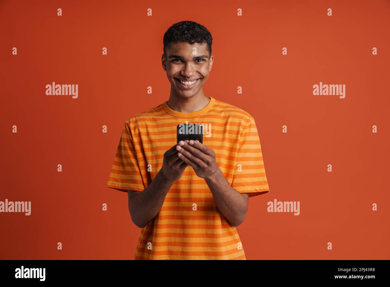 Ein junger, gut aussehender afrikaner mit Piercings, der sein Handy hielt und in die Kamera schaute, während er über einem isolierten orangefarbenen Hintergrund stand Stockfoto