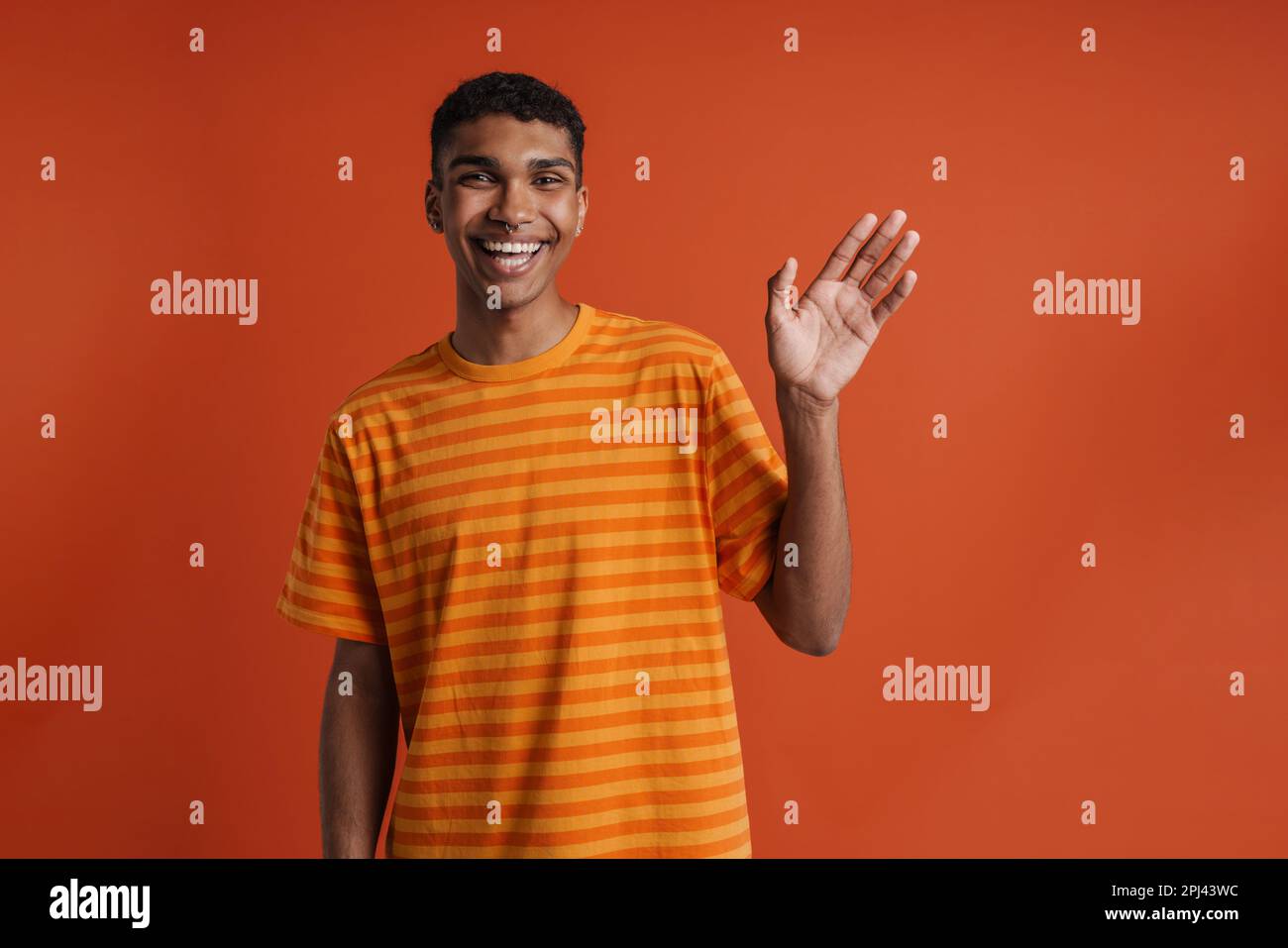 Junger, gutaussehender, glücklicher afrikanischer Mann mit Piercing, der winkte und in die Kamera schaute, während er über einem isolierten orangefarbenen Hintergrund stand Stockfoto