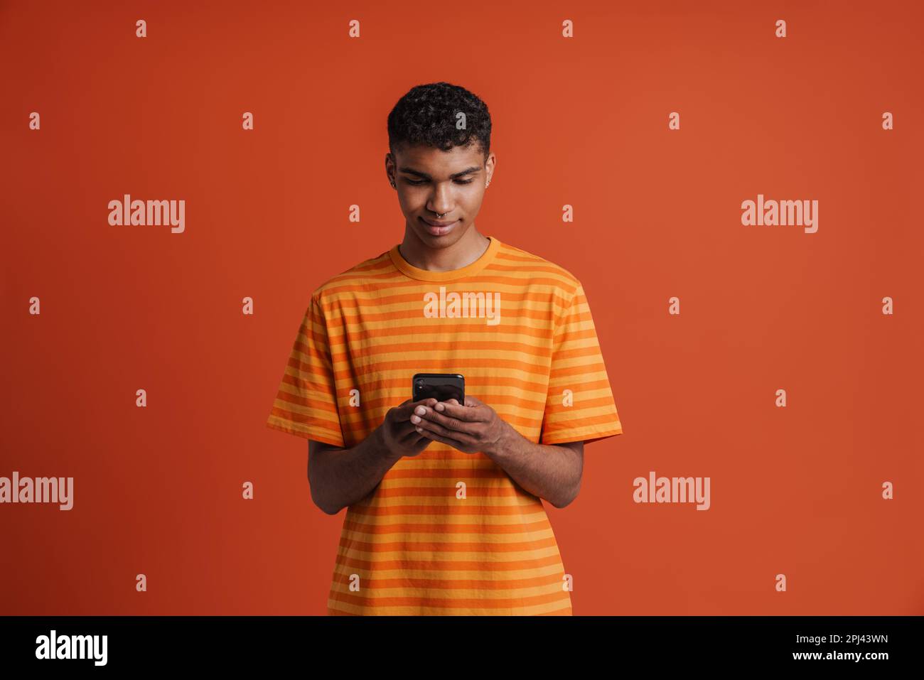 Ein junger, gut aussehender, ruhiger afrikaner mit Piercings, der sein Handy hält und darauf schaut, während er über einem isolierten orangefarbenen Hintergrund steht Stockfoto