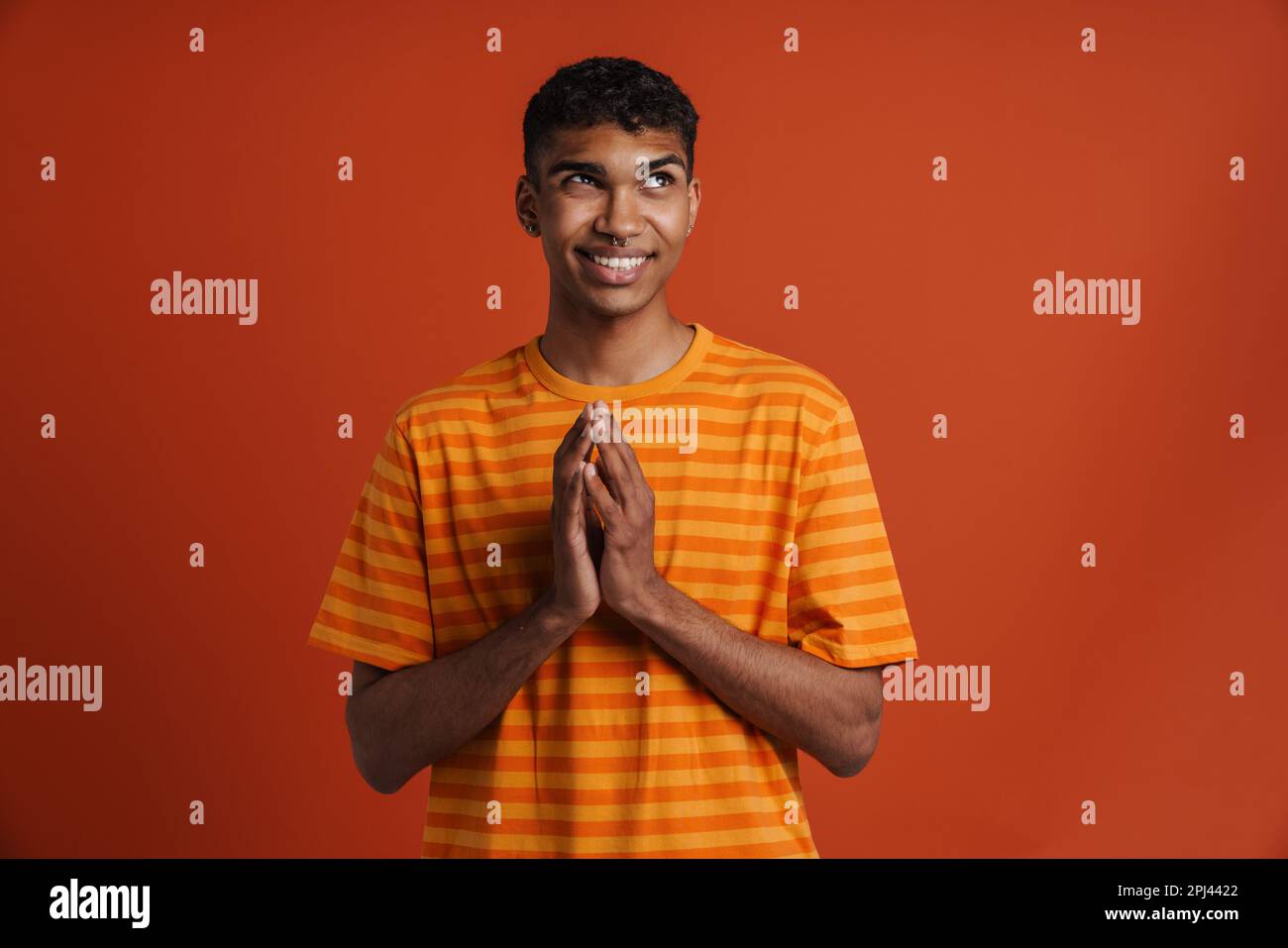 Junger schwarzer Mann mit einem stechenden Lächeln, während er Gebetsgesten isoliert vor rotem Hintergrund macht Stockfoto
