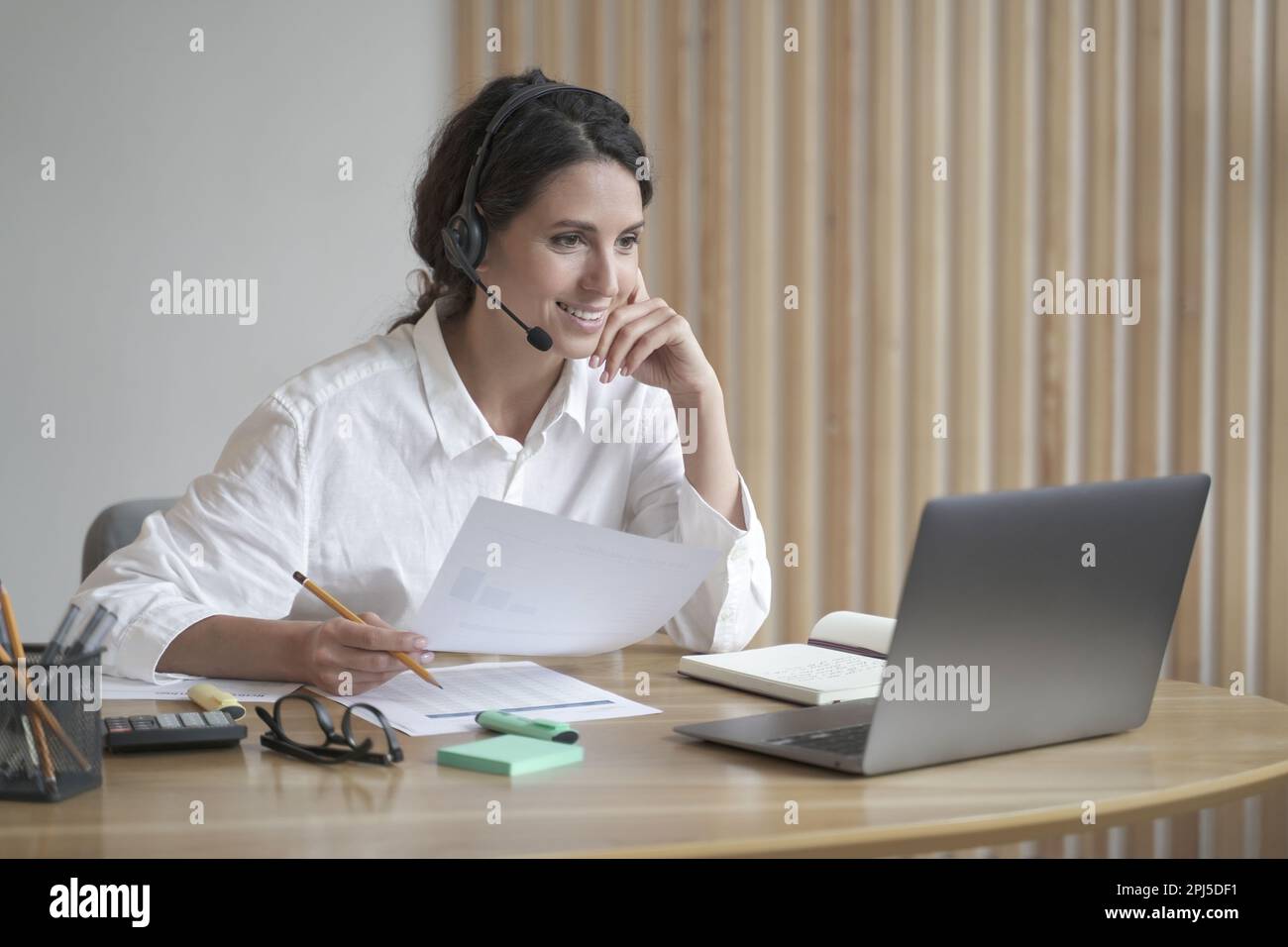 Positive junge Callcenter-Agentin in einer schnurlosen Headset-Beratungskunde während der Arbeit im Büro, hispanische Lehrerinnen als Tutor geben weniger online Stockfoto