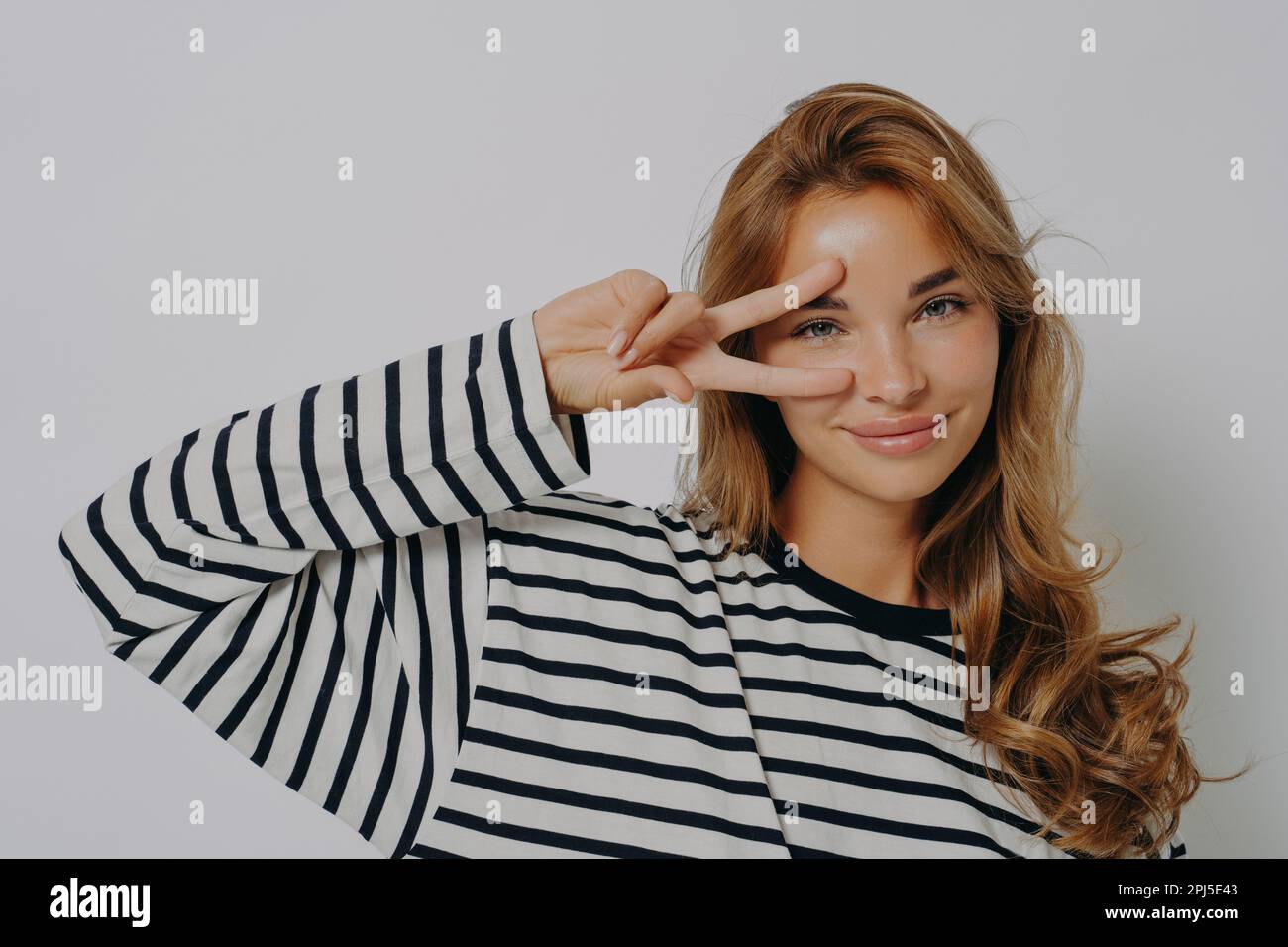 Glückliche junge europäische Frau macht Friedensdisco-Geste über Auge V-Schild trägt lässigen gestreiften Pullover minimales Make-up steht drinnen gegen graue Rückseite des Studios Stockfoto