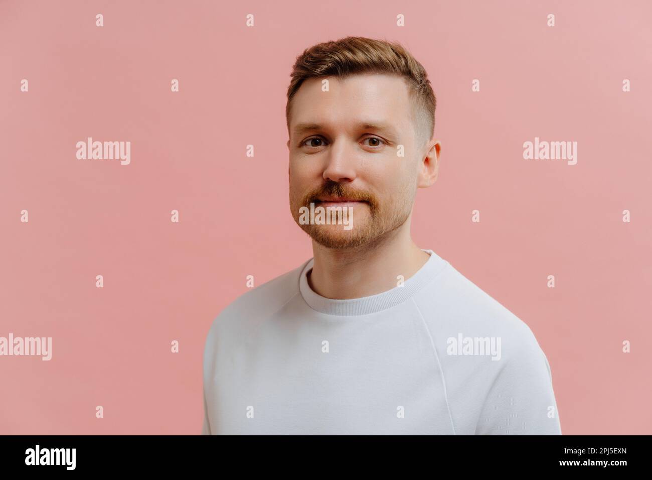 Ein wahrhaft gutaussehender europäischer Mann mit drei-Tage-Bart sieht direkt in die Kamera. Er trägt ein legeres weißes T-Shirt und verfügt über ruhige Gesichtsausdrücke vor pinkfarbenem Backgr Stockfoto