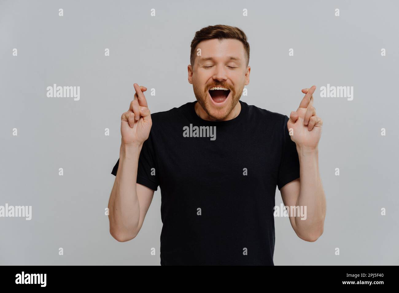 Optimistisch aufgeregter europäischer Mann mit Bart, der sicher ist, dass der Wunsch wahr werden wird, drückt die Daumen und wartet auf positive Ergebnisse hält den Mund offen und die Augen geschlossen Stockfoto