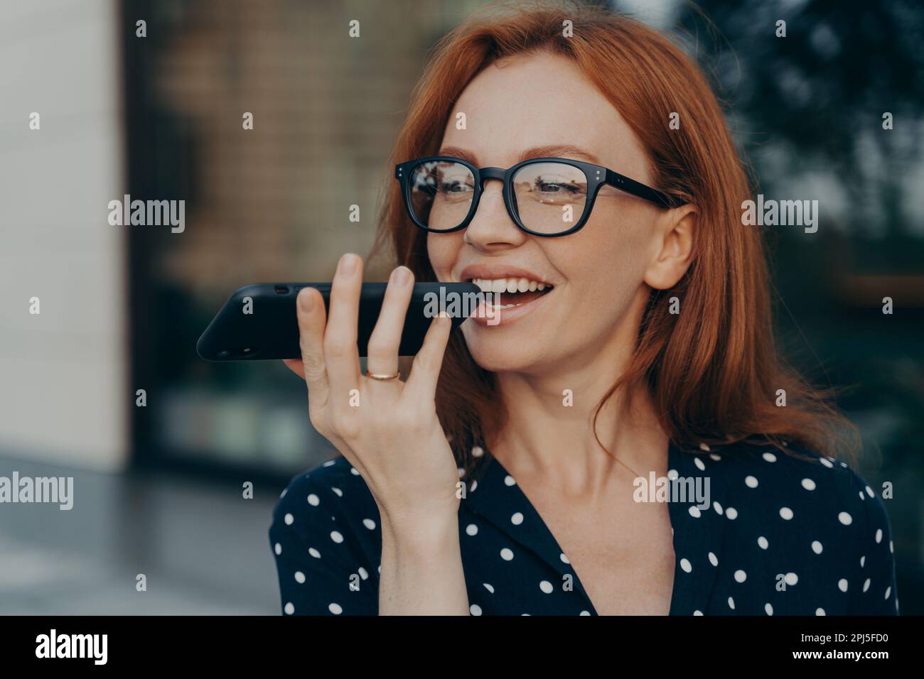 Nahaufnahme einer jungen, lächelnden rothaarigen Geschäftsfrau, die im Freien mit ihrem Mobiltelefon steht und einen virtuellen digitalen Sprachassistenten verwendet, glückliche Frau Stockfoto