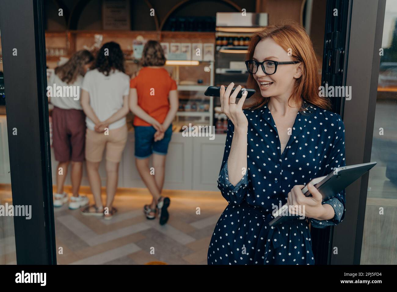 Die hübsche rothaarige Frau nimmt Audionachrichten über das Handy auf. Sie trägt einen Laptop und ein Tagebuch sieht vor dem Hintergrund des Ladens aus und trägt Polka Dotte Stockfoto