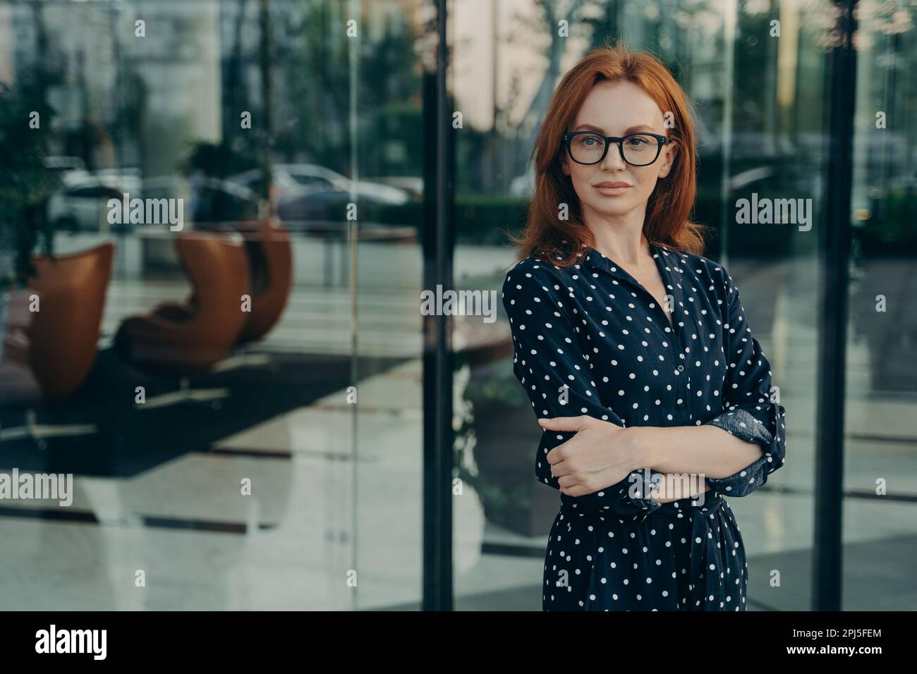 Selbstsichere Regisseurin mit rotem Haar angenehmes Aussehen in elegantem Kleid Brille Posen in der Nähe des Bürogebäudes wartet auf Partner gehen Stockfoto