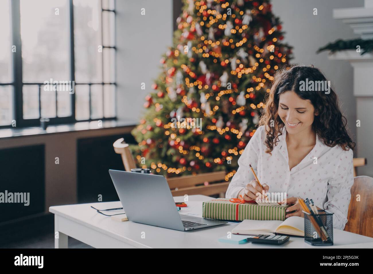 Junge, zufriedene europäische Büroangestellte schrieb einen weihnachtsbrief, Grußkarte mit Urlaubswünschen, während sie mit ihrem Laptop am Arbeitsplatz saß und Stockfoto
