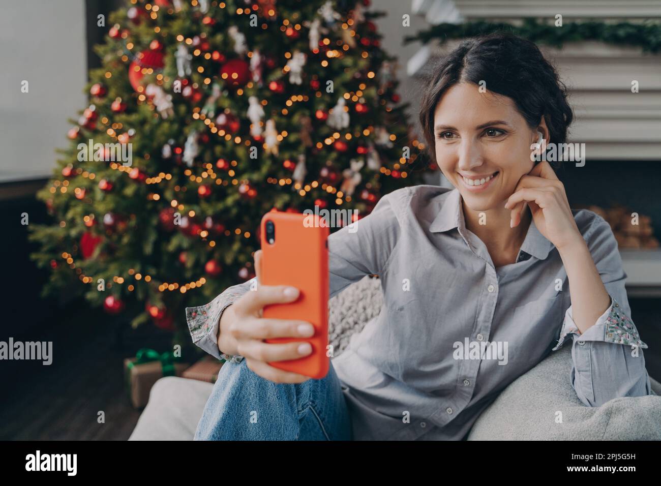 Fröhliche junge spanierin, die die weihnachtszeit genießt und Selfie macht, während sie auf dem bequemen Sofa neben dem festlichen, bunten Weihnachtsbaum sitzt. Angenehmes Blo Stockfoto