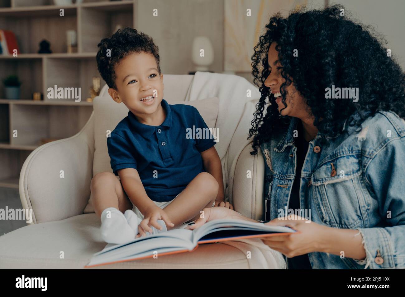 Leidenschaftliche, positive afro-amerikanische Mutter, die ihrem süßen, aufgeregten kleinen Sohn lustige Geschichten vorliest, zusammen lacht und Zeit damit verbringt, über A zu lernen Stockfoto