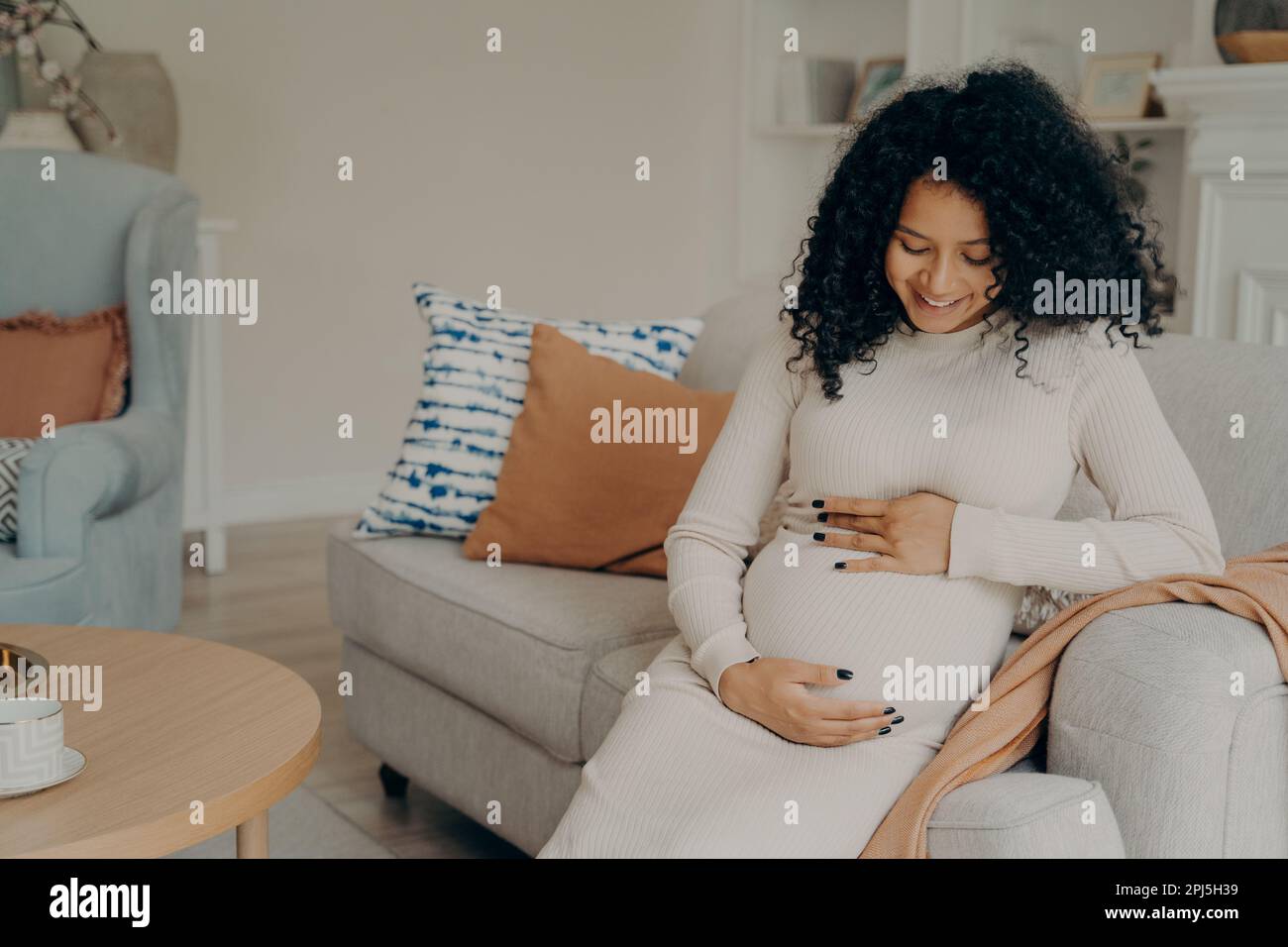 Glückliche junge, schwangere afro-amerikanische Dame in weißem Kleid mit langen lockigen Haaren, die ihren Bauch mit einem sanften Lächeln ansieht und eine kleine Pause von der Hausarbeit macht Stockfoto