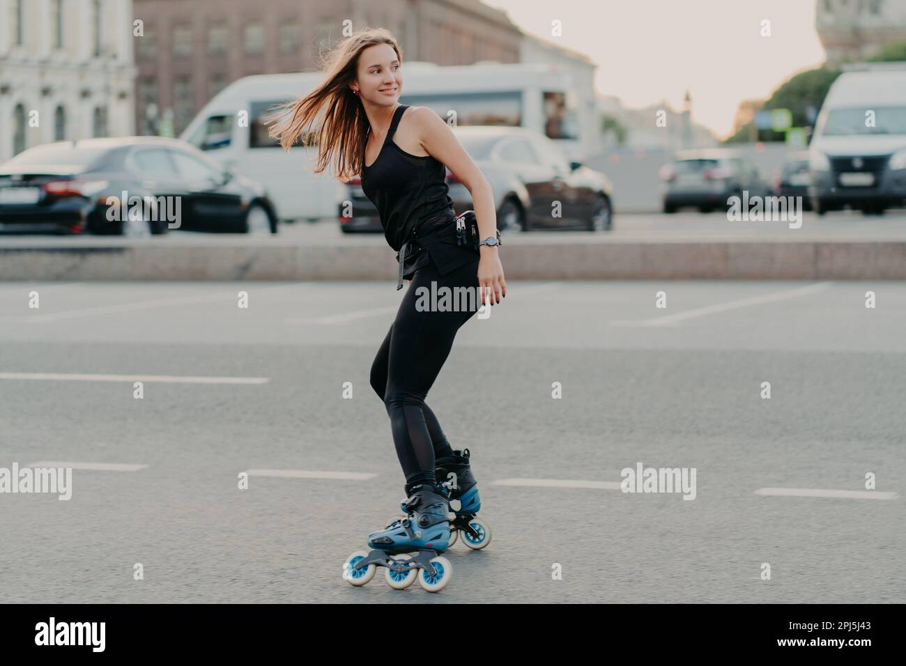 Seitlich von einer aktiven jungen Frau in guter körperlicher Form aufgenommen Rollerblades entlang der Straßen der Stadt sieht glücklich dahinter trägt schwarze Sportkleidung führt Gesundheit Stockfoto