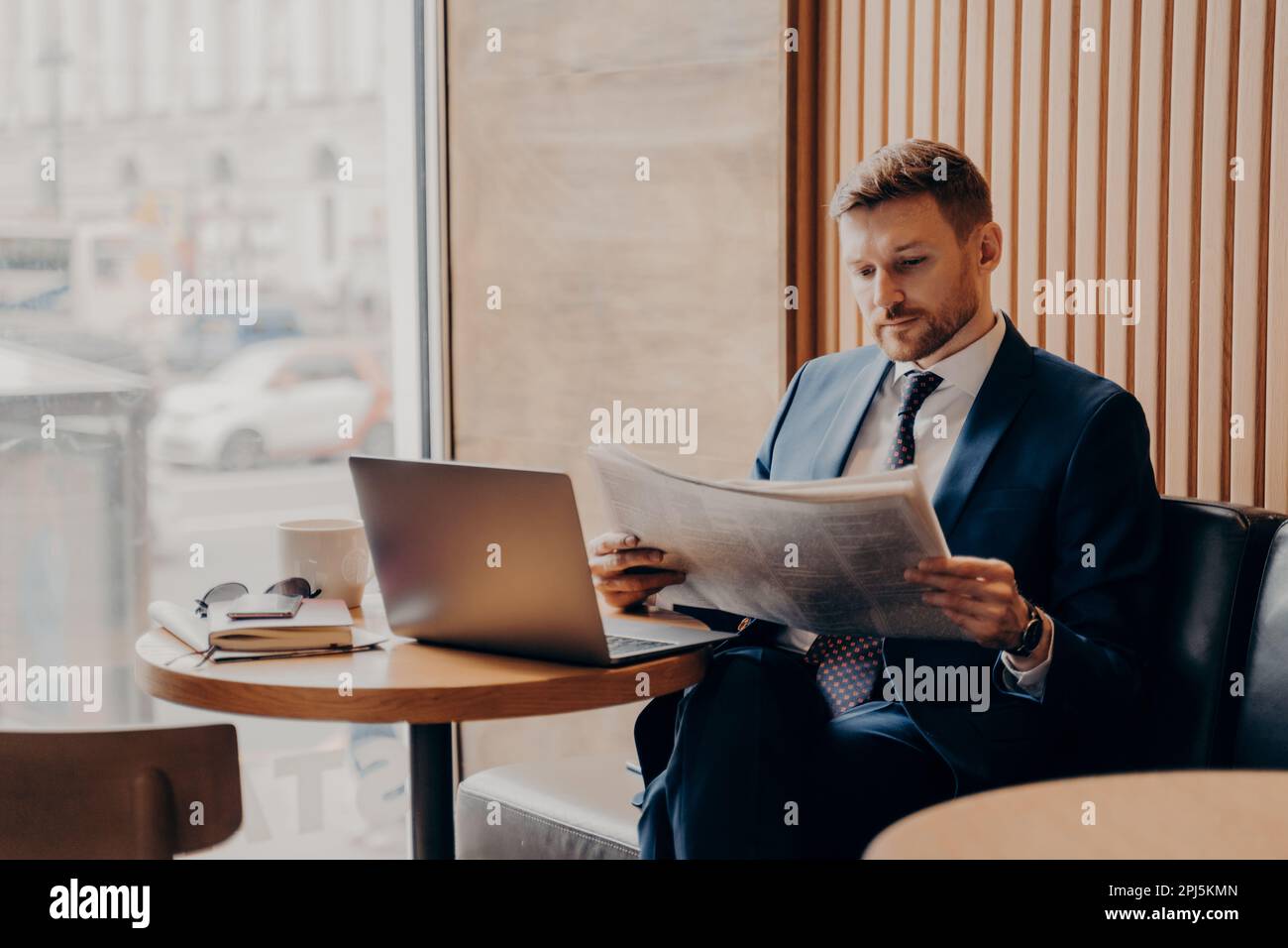 Konzentrierter männlicher Geschäftsangestellter in blauem formellen Anzug, sitzt in der Ecke des Cafés neben dem großen Fenster, liest Zeitung aus nächster Nähe, Arbeitslappen öffnen Stockfoto