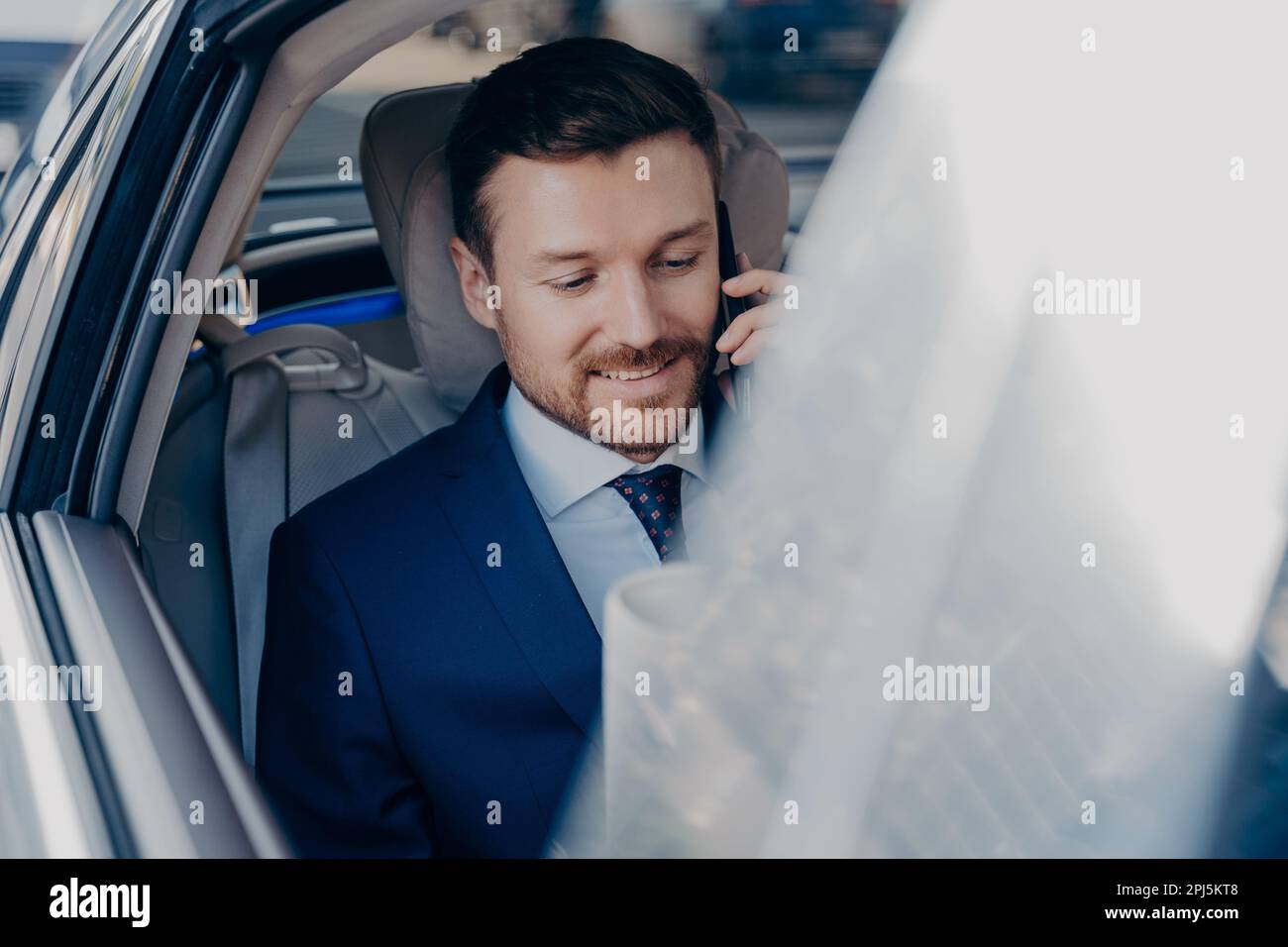 Junger positiver Geschäftsmann fährt im Auto auf dem Beifahrersitz in das Büro im Stadtzentrum, spricht mit fokussiertem Aussehen und lächelndem Smartphone. Seitenbild von Stockfoto