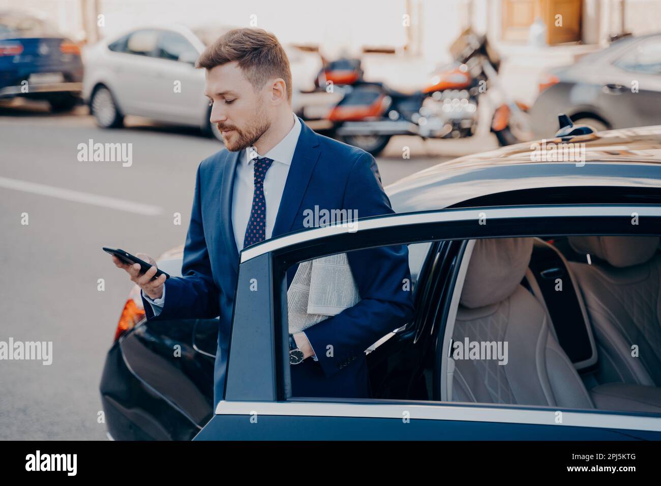 Erfolgreicher junger, gut gepflegter Manager in formellem, stilvollem Anzug, der aus dem Auto der Premiumklasse aussteigt, am Arbeitsplatz ankommt, während er nach verpassten Stockfoto