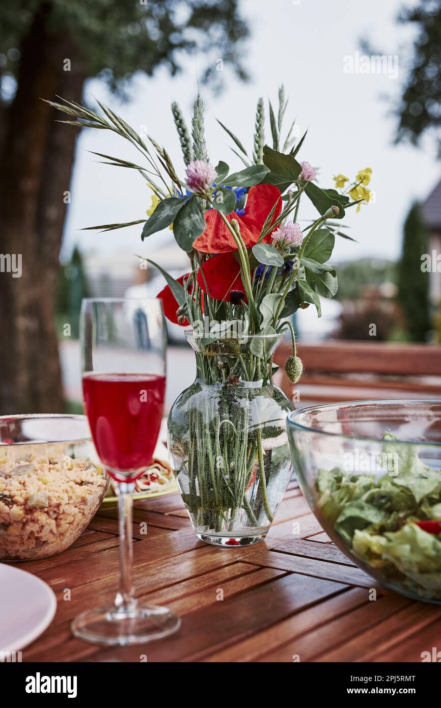 Abendessen in einem Apfelgarten auf Holztisch mit Salaten und Wein mit Blumen dekoriert. Nahaufnahme des Tisches mit Essen für Familienessen vorbereitet Stockfoto