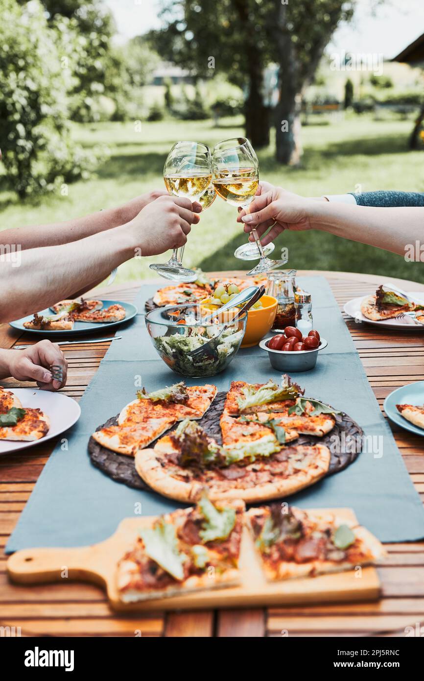 Freunde, die während des Picknick-Abendessens im Freien in einem heimischen Garten anstoßen. Nahaufnahme von Leuten, die Weingläser mit Weißwein über den Tisch mit piz halten Stockfoto
