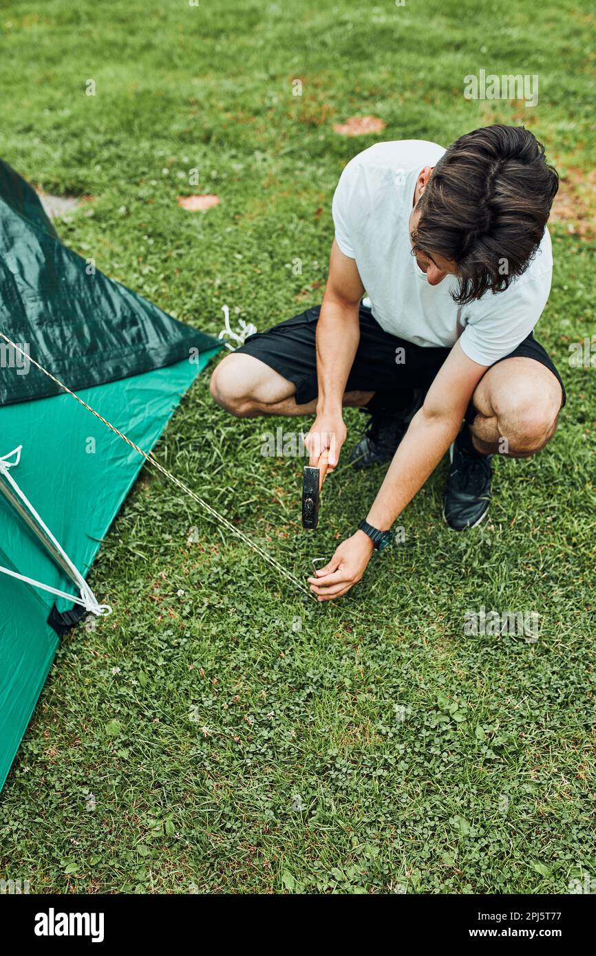 Junger Mann, der während der Sommerurlaubsreise ein Zelt auf dem Campingplatz aufstellt. Teenager, der die Spieße mit Hammer auf Grasboden legt Stockfoto