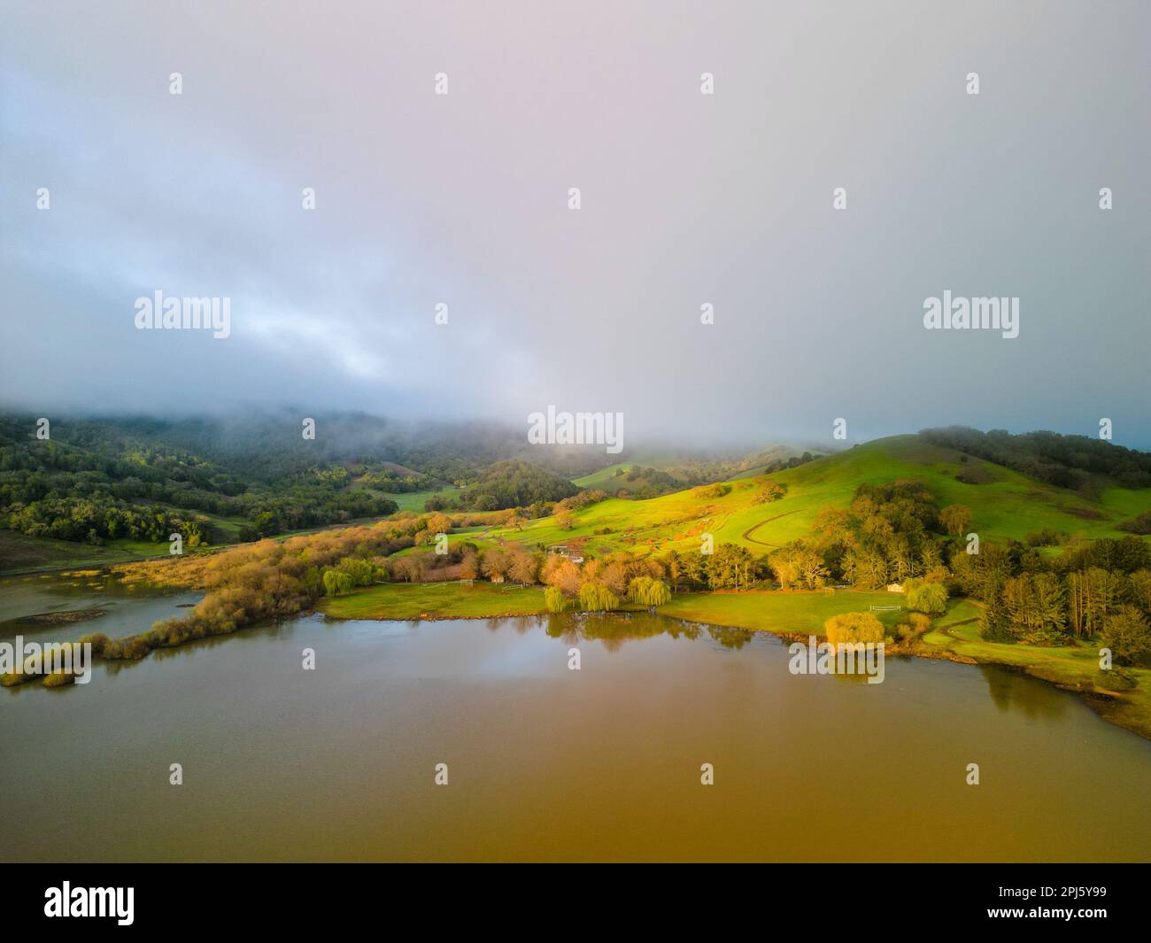 Am frühen Morgen trifft die Sonne auf grüne Hügel am See unter niedriger Nebelschicht Stockfoto