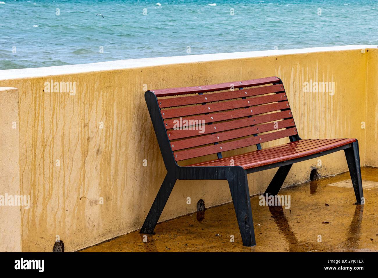 Leere hölzerne Bank, nass von Meerwasser neben einem Zaun an der Strandpromenade, Meer im Hintergrund, blaues Wasser ohne Wellen, ruhiger und bewölkter Tag in La Paz, Stockfoto