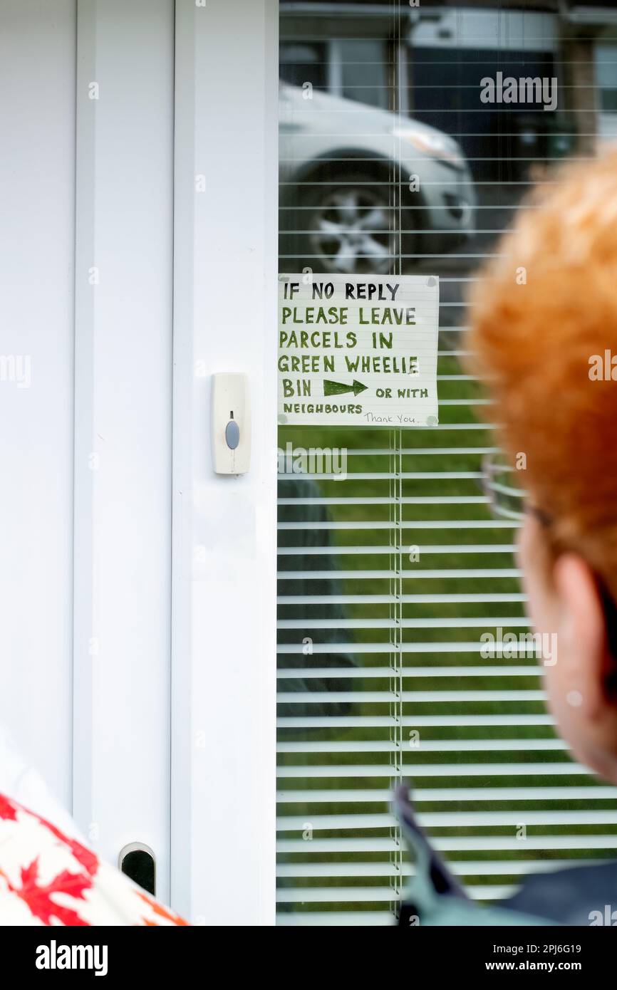 Ein Lieferkurier liest einen Lieferschein im Fenster der Lieferadresse. In der Notiz werden Sie aufgefordert, Pakete an einem sicheren Ort zu lassen, wenn keine Antwort erfolgt Stockfoto