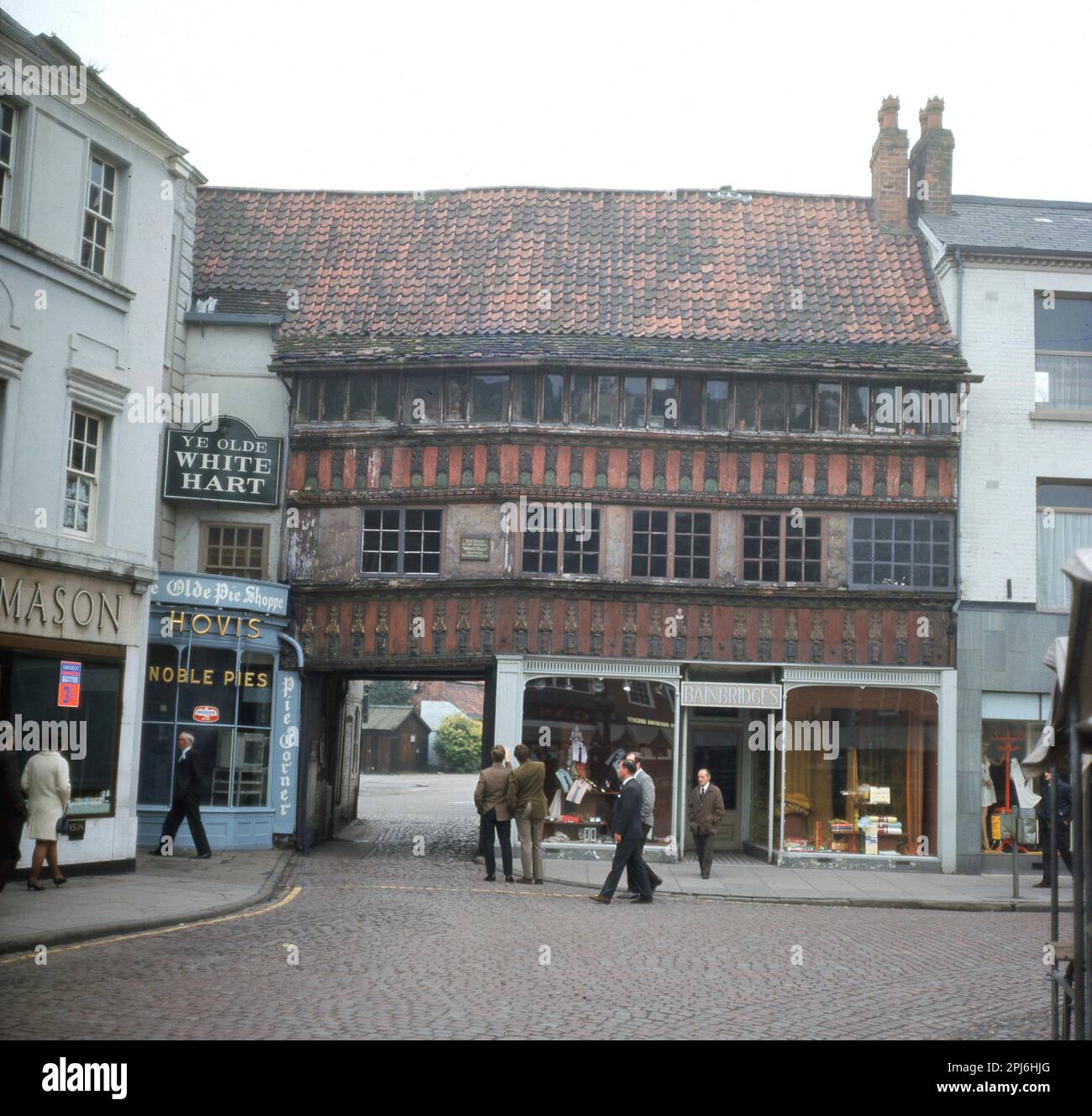 Etwa Ende 1960er, historischer Blick von außen aus dieser Ära, auf einer gepflasterten Straße, zwei Männer blicken auf ein altes Gebäude, Ye Olde White Hart an der südöstlichen Ecke des Market Place, Newwark on Trent, England, Großbritannien. Dieses alte Gasthaus mit gewölbtem Eingang stammt aus dem 14. Jahrhundert. Auf der linken Seite, neben ihr, ihr Olde Pie Shoppe, der Noble Pies verkauft. Unter dem alten Gasthaus, einem Schuppen, Bainbridges. Shoppen Sie links von Ye Olde White Hart, Mason, mit einem Werbeposter für Kangaroo Butter, 3/- ( 3 Schilling) im Fenster, Stockfoto