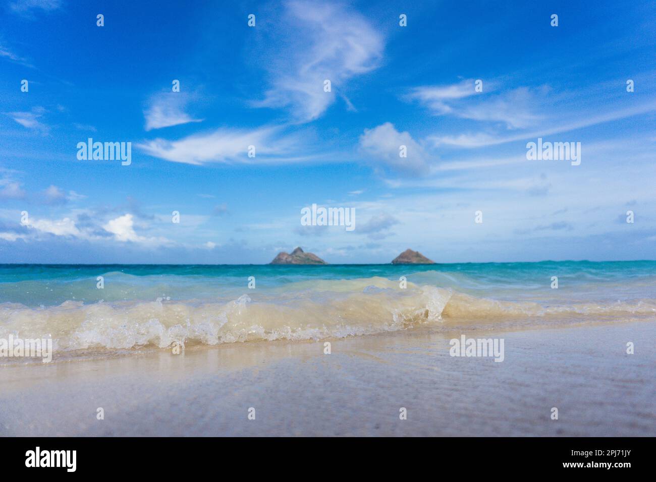 Niedriger Blickwinkel auf tropische Wellen, die sich bis zum Sandstrand in Hawaii mit kleinen Inseln in der Ferne und blauem Himmel oben waschen Stockfoto
