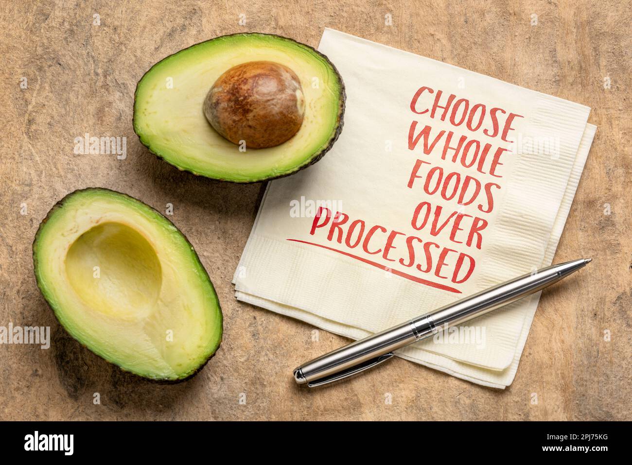 Entscheiden Sie sich für ganze Lebensmittel über verarbeitete - gesundes Ernährungs- und Lifestyle-Konzept, Erinnerungsworte handgeschrieben auf einer Serviette mit geschnittenen Avocadofrüchten Stockfoto