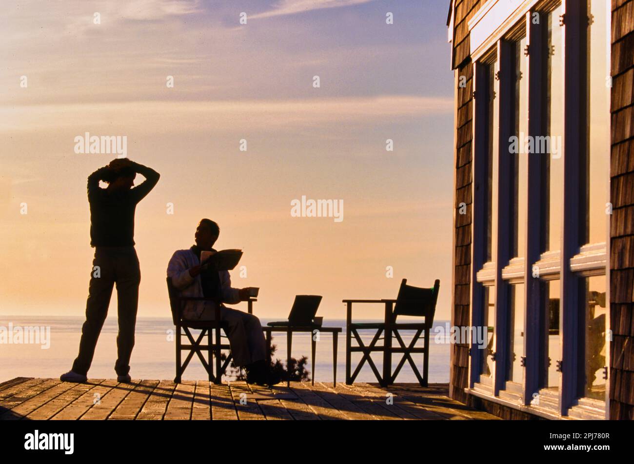 Eine Silhouette von einem Paar, das auf der Terrasse seines Ferienhauses mit Blick auf den Ozean bei Sonnenuntergang sitzt Stockfoto