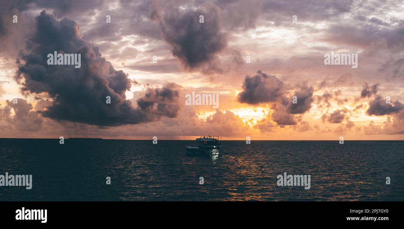 Ein atemberaubender Sonnenuntergang mit unauffälligem Panoramablick auf ein Boot im Meer. Die orangefarbene Sonne ist teilweise hinter violetten und rosafarbenen Wolken verborgen; die Silhouette des Boots Stockfoto