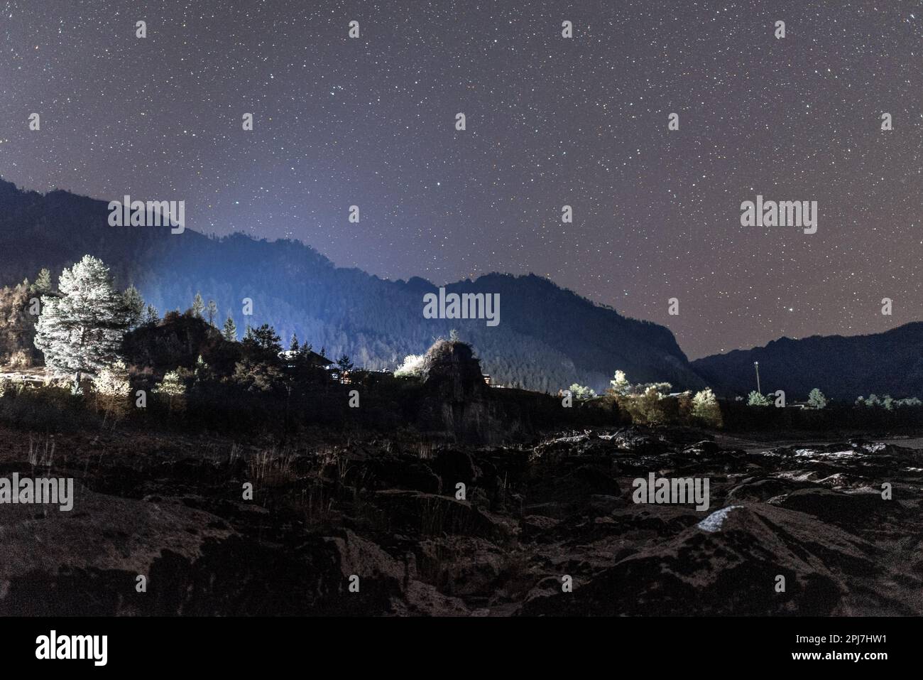 Das Steinufer des Flusses Katun im Schatten bei Nacht nahe der Silhouette eines Berges, der durch das Licht der anderen Häuser unter den Sternen beleuchtet wird Stockfoto