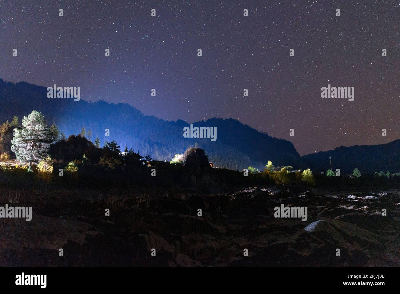 Das Steinufer des Flusses Katun im Schatten bei Nacht nahe der Silhouette eines Berges, der durch das Licht der anderen Häuser unter den Sternen beleuchtet wird Stockfoto