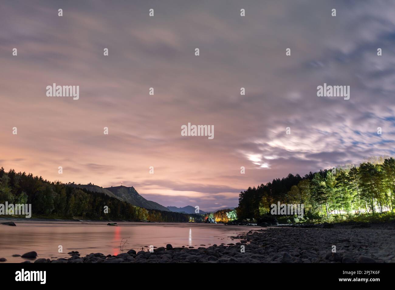 Helle Nacht mit Wolken am Himmel in der Nähe des Bergflusses Katun mit einem roten Radiosender auf dem Felsen und dem Licht der Raststätten im Wald in diesem Stockfoto