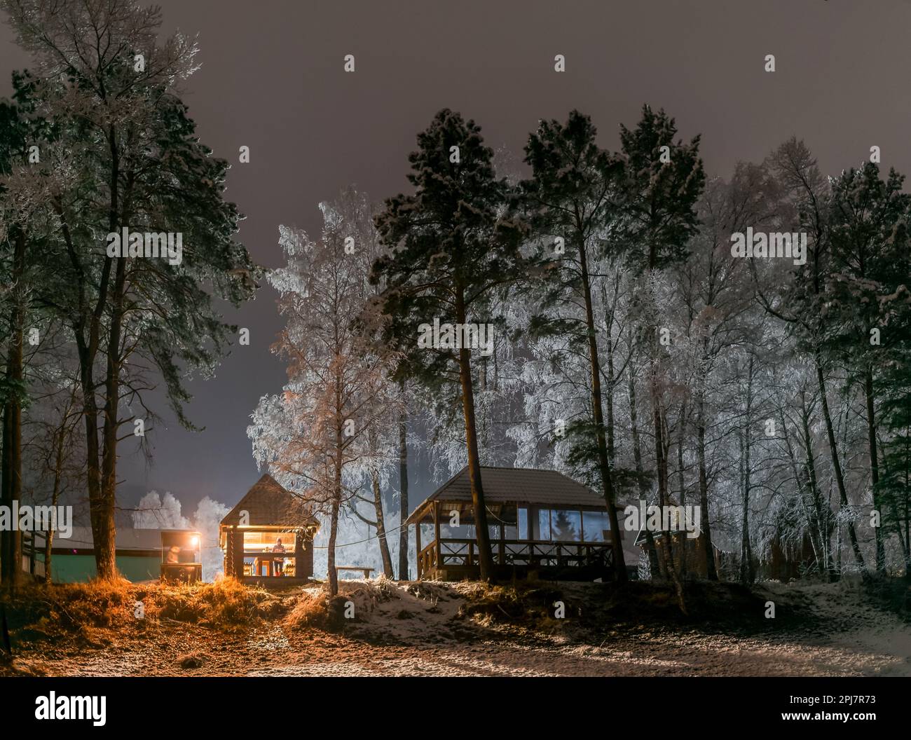 Ferienhaus am Ufer mit zwei Frauen, die sich nachts im Schnee unter Birkenbäumen ausruhen, mit einem Grillfeuer in Altai in Sibirien im Dunkeln. Stockfoto