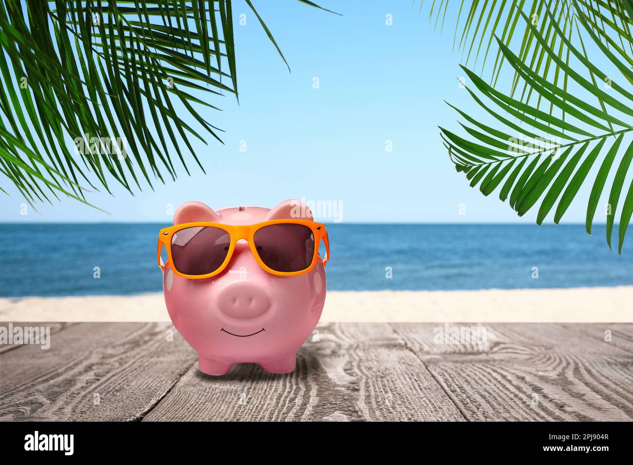 Sparen Geld für den Sommerurlaub. Sparschwein mit Sonnenbrille auf Holzfläche in der Nähe von Sandstrand und Meer Stockfoto