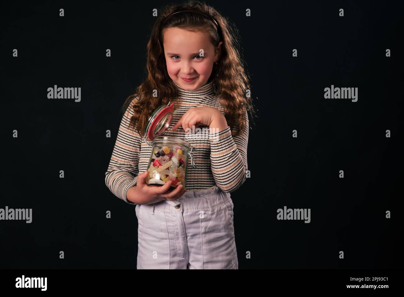 8-jähriges Mädchen hält ein Glasgefäß mit bunten Süßigkeiten, zeigt mit einem unartigen Lächeln auf Süßigkeiten, während es in die Kamera schaut. Isoliert auf dunklem Hintergrund Stockfoto