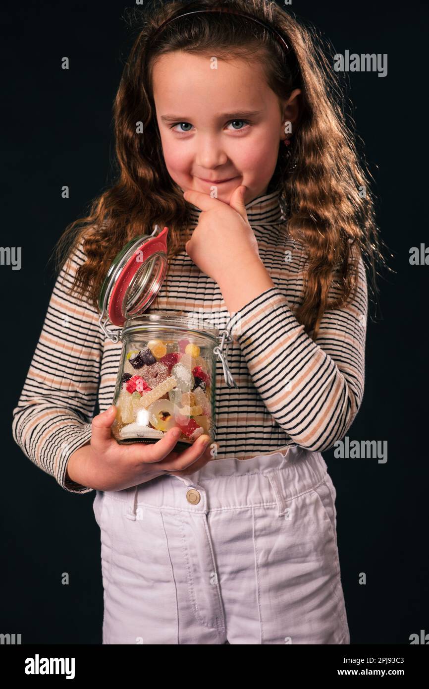 8-jähriges Mädchen hält Glasbecher mit bunten Süßigkeiten, hält Hände am Kinn und schaut in die Kamera. Essen oder nicht essen. Dunkler Hintergrund Stockfoto
