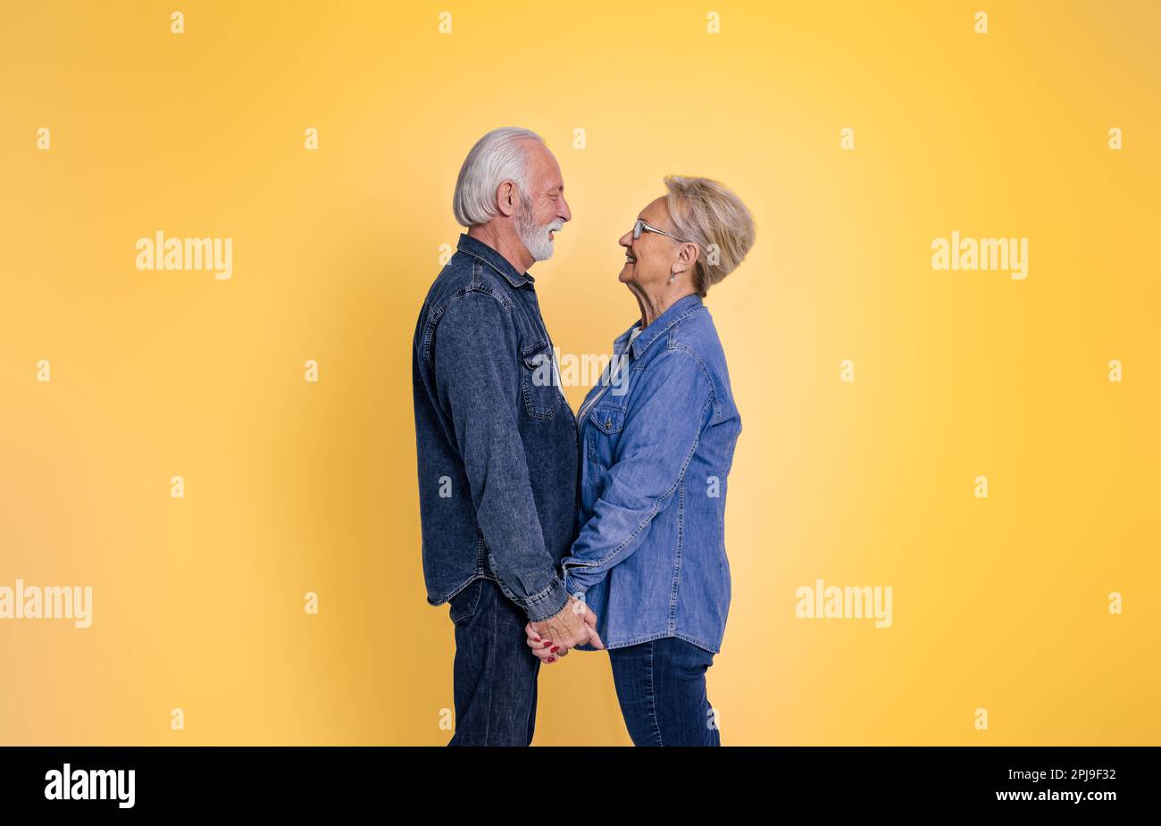 Seitenansicht eines liebevollen Seniorenmannes und einer liebevollen Seniorenfrau, die Händchen hält und sich romantisch anschaut, während sie isoliert auf gelbem Hintergrund stehen Stockfoto