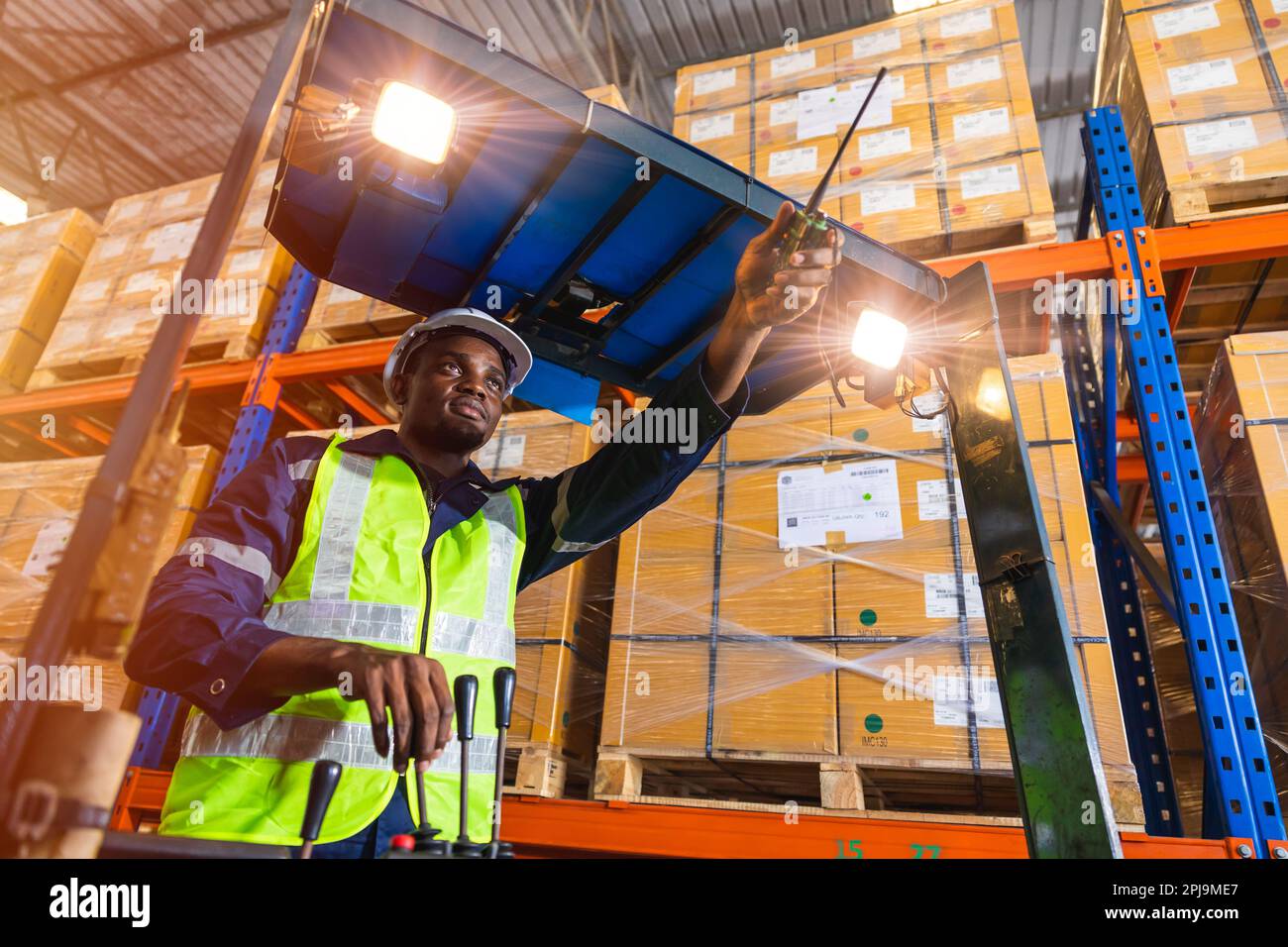 Afrikanischer schwarzer männlicher Angestellter, der die Kontrolle über das Verladen ausübt, betreibt einen Volkslift in der Lagerbestandsfabrik Stockfoto