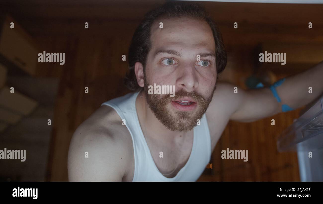 Hungriger Mann mit Sportler, Bart und langen Haaren findet kein Essen im Kühlschrank Stockfoto